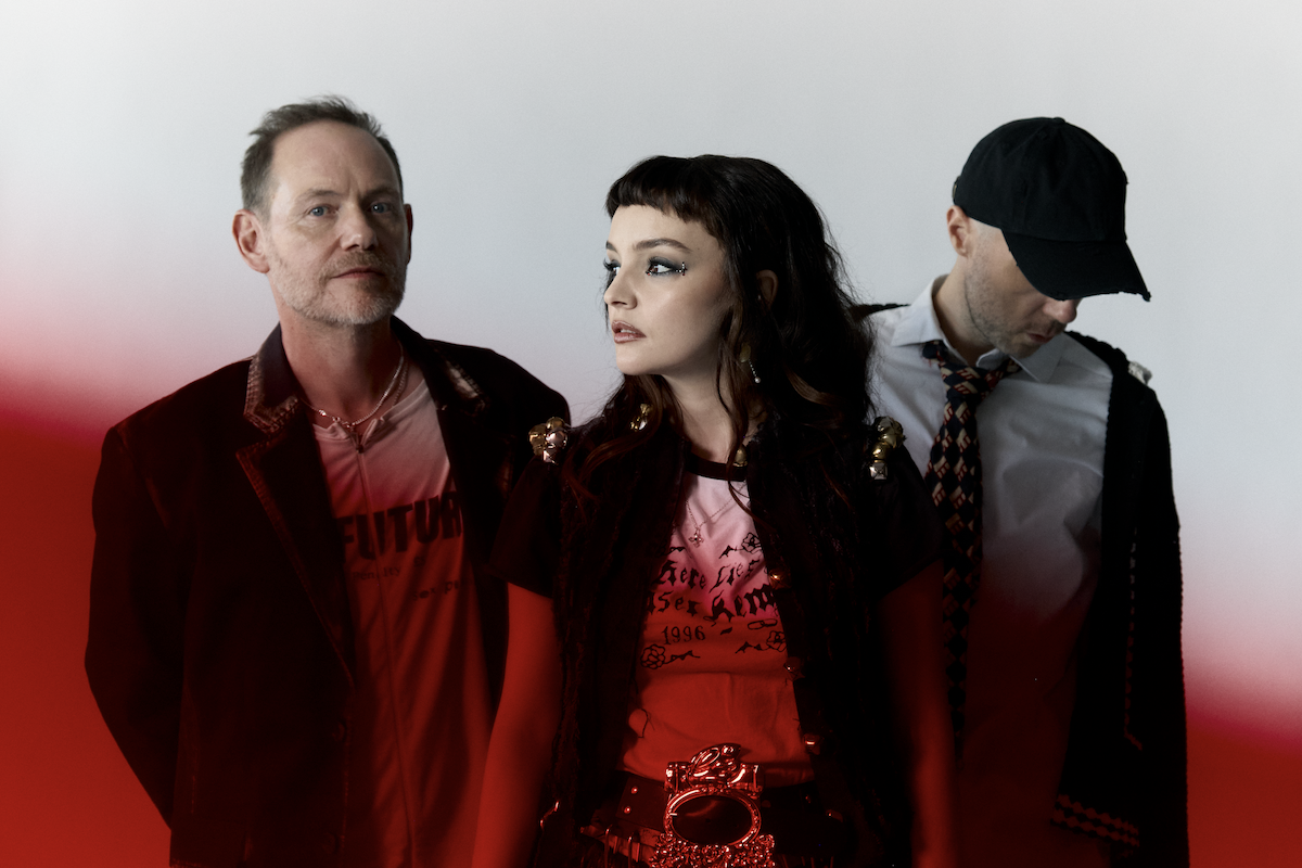 Scottish synth-pop band CHVRCHES featuring Lauren Mayberry, Iain Cook, and Martin Doherty in a studio portrait against red background.